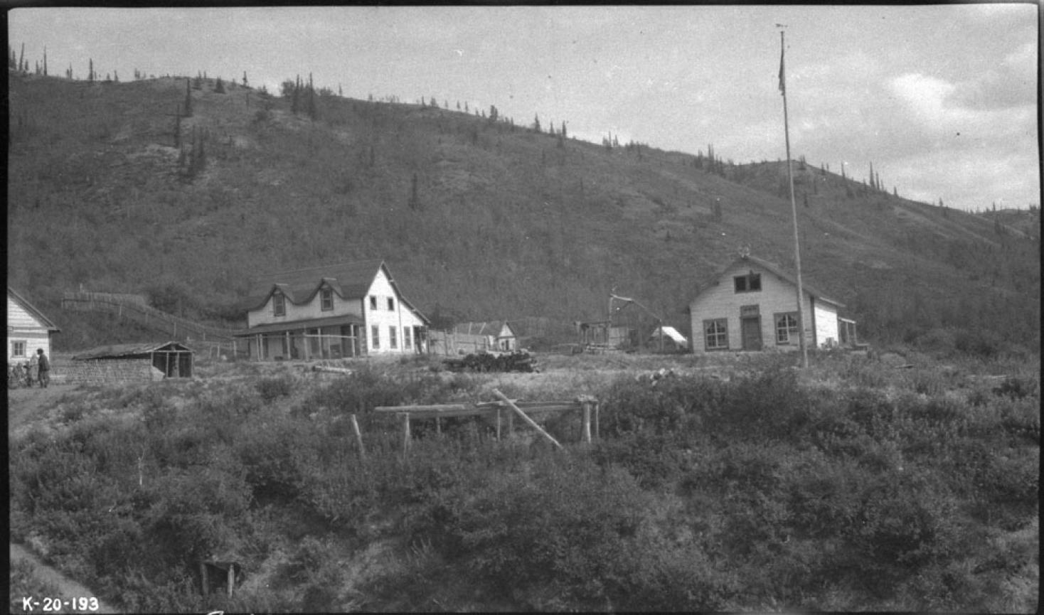 Archival black and white photo of buildings on a grassy area with a hilly mountain behind them.